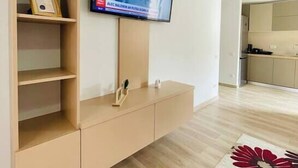 Un salon moderne avec un téléviseur à écran plat fixé au mur, un meuble en bois doté d’étagères ouvertes et un coin cuisine visible à travers une porte ouverte.