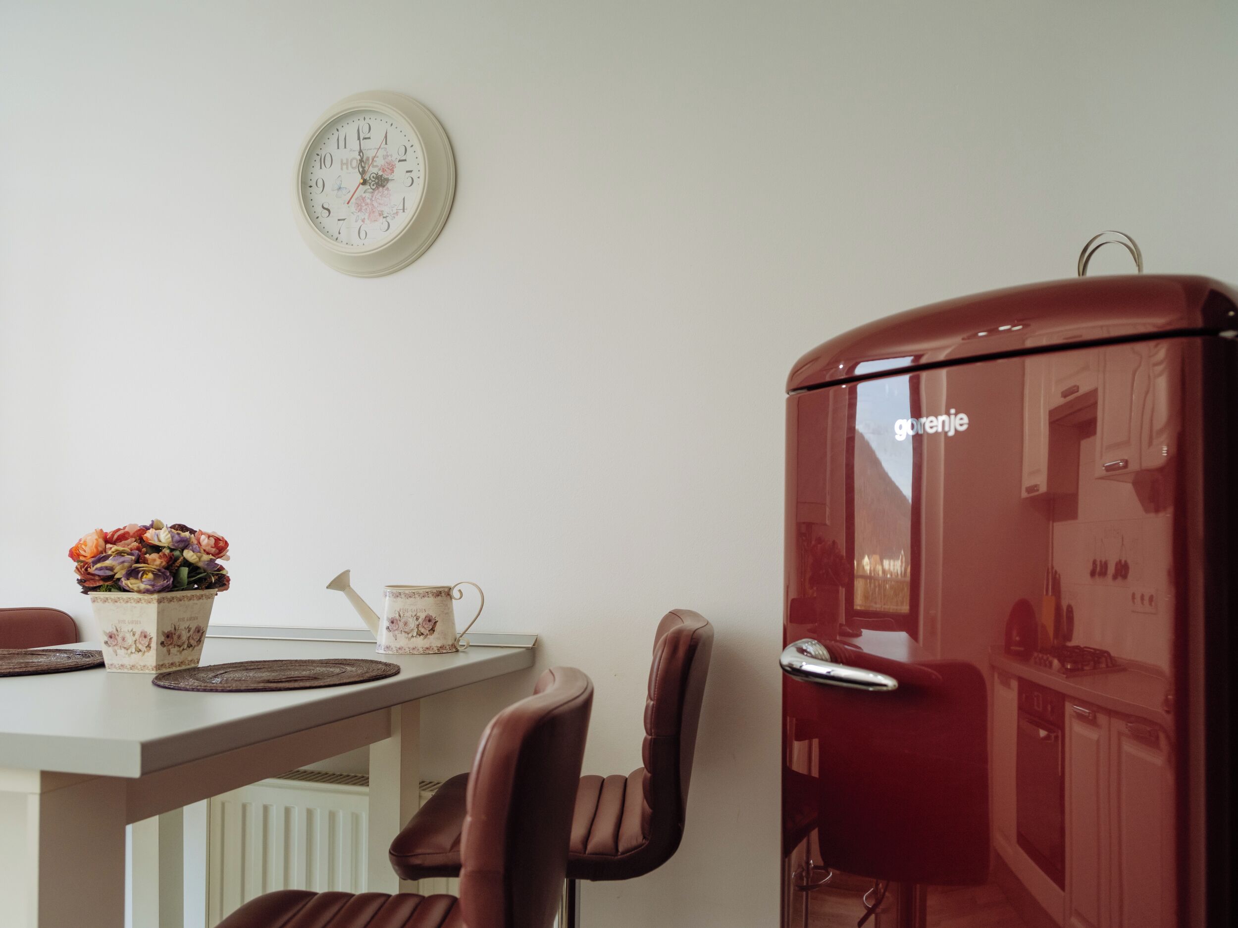 A kitchen with a red refrigerator, a dining table with chairs, a floral vase, and a wall clock.