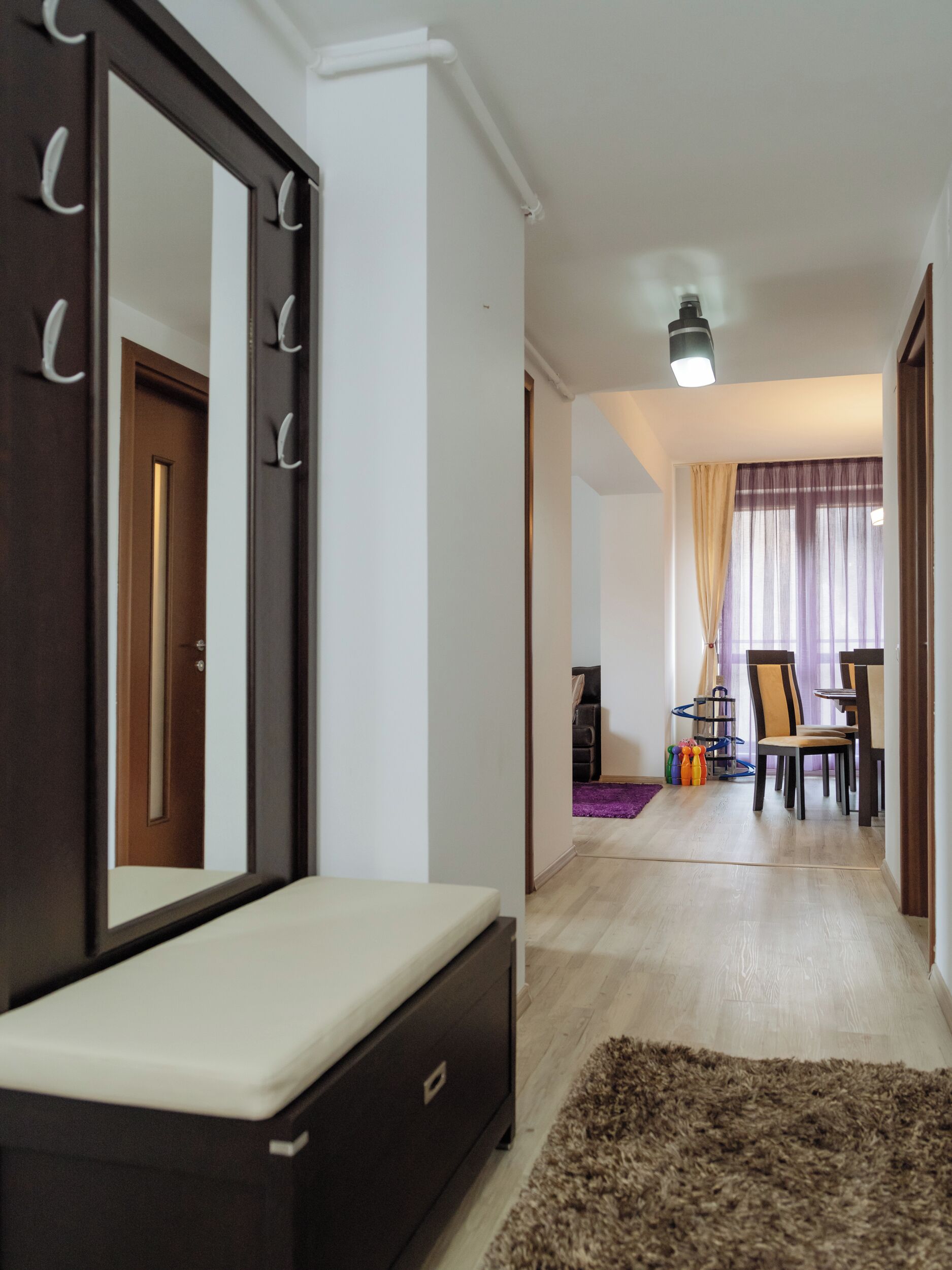A room with a large mirror, a wooden door, a bench, a rug, a dining area with chairs and a table, and a ceiling light.