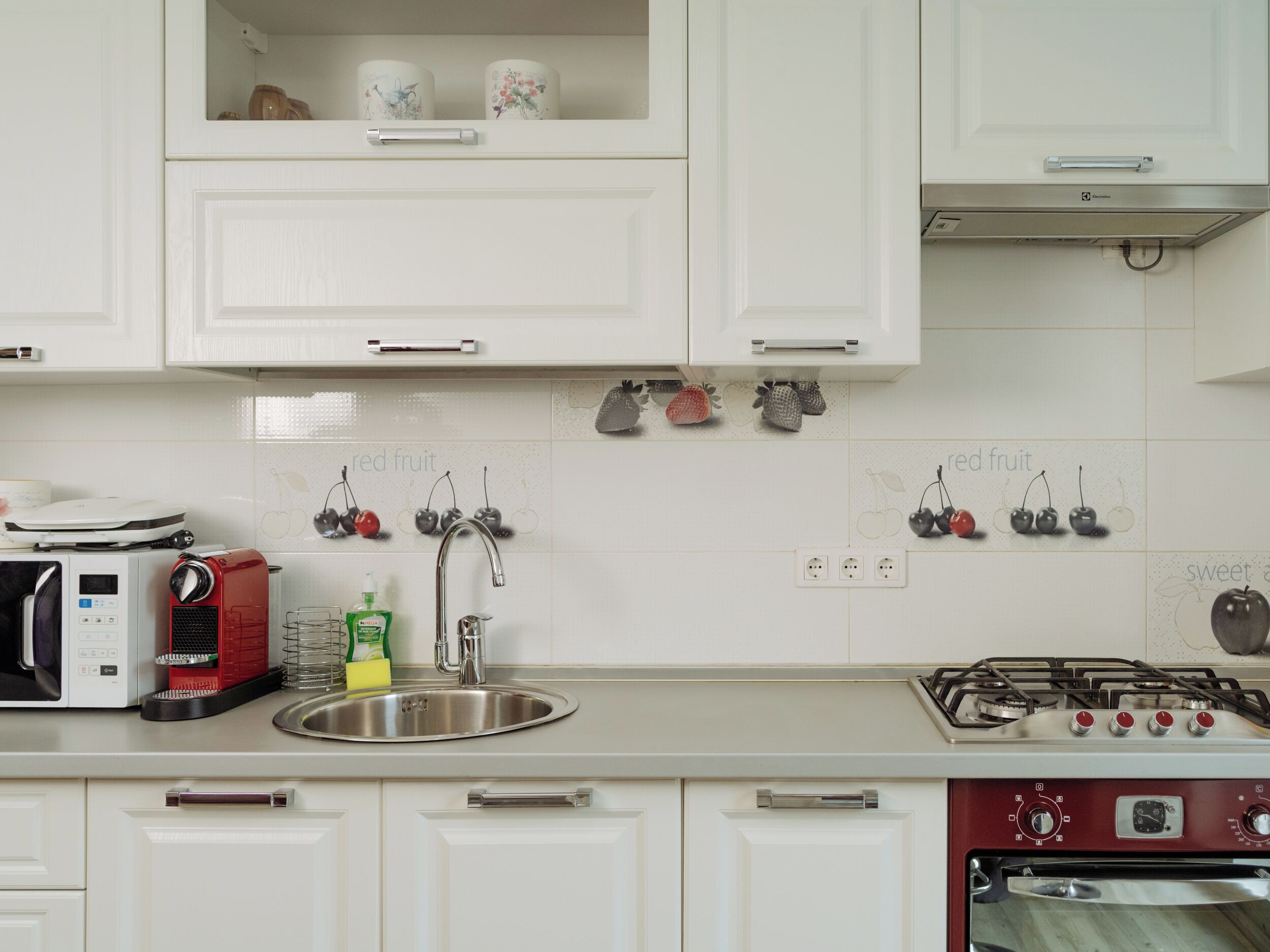 Une cuisine avec des meubles blancs, un évier en acier inoxydable, une cuisinière à gaz rouge et un four à micro-ondes.