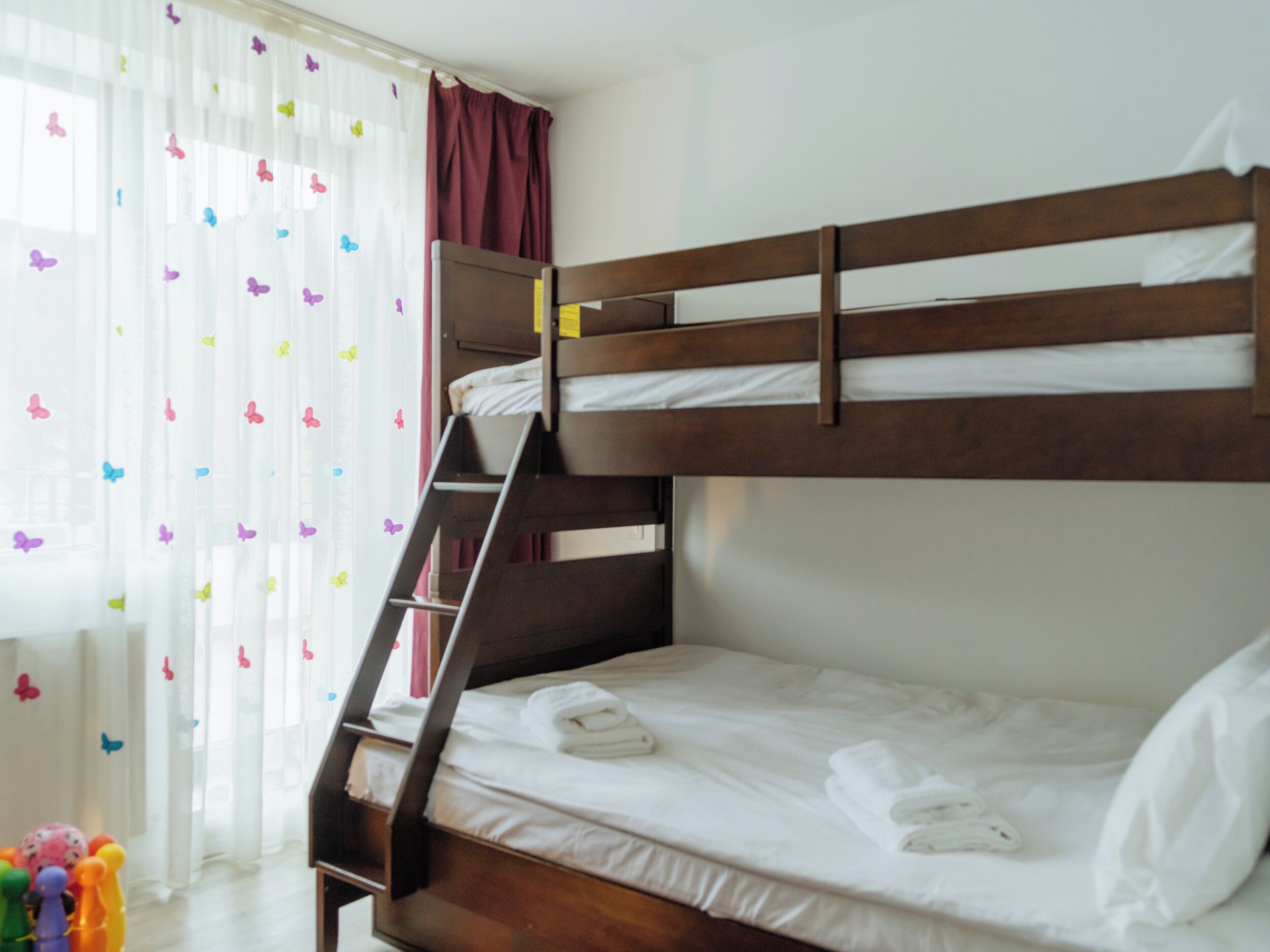 Een stapelbed met houten frame, witte beddengoed en een ladder. Er is een raam met dunne gordijnen en vlinderstickers.