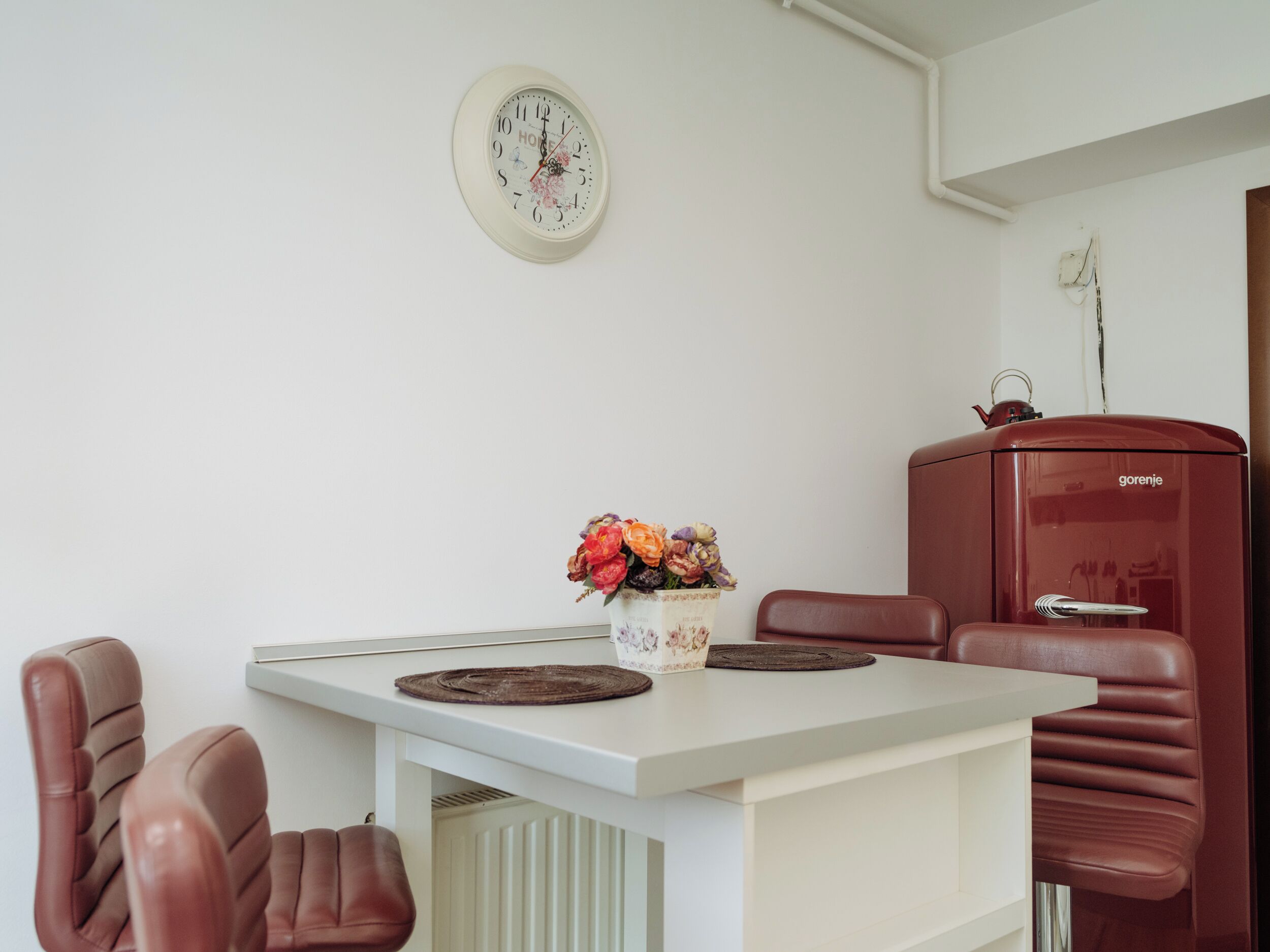 Un coin repas avec une table blanche, des chaises en cuir marron, un vase de fleurs et un appareil électroménager rouge accroché au mur.