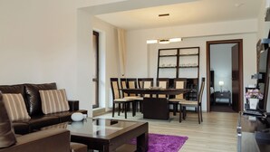 Una sala de estar moderna con un sofá de cuero marrón oscuro, una mesa de centro de vidrio y un tapete morado. Hay una zona de comedor con una mesa y sillas de madera oscura, y un pasillo que conduce a un dormitorio.