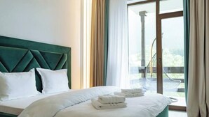 Quarto de hotel com cama grande, cabeceira verde, roupa de cama branca e vista para uma varanda com uma cadeira.