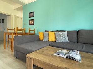 A living room with a grey sectional sofa, wooden chairs, a coffee table with a magazine, and two framed pictures on a teal wall.