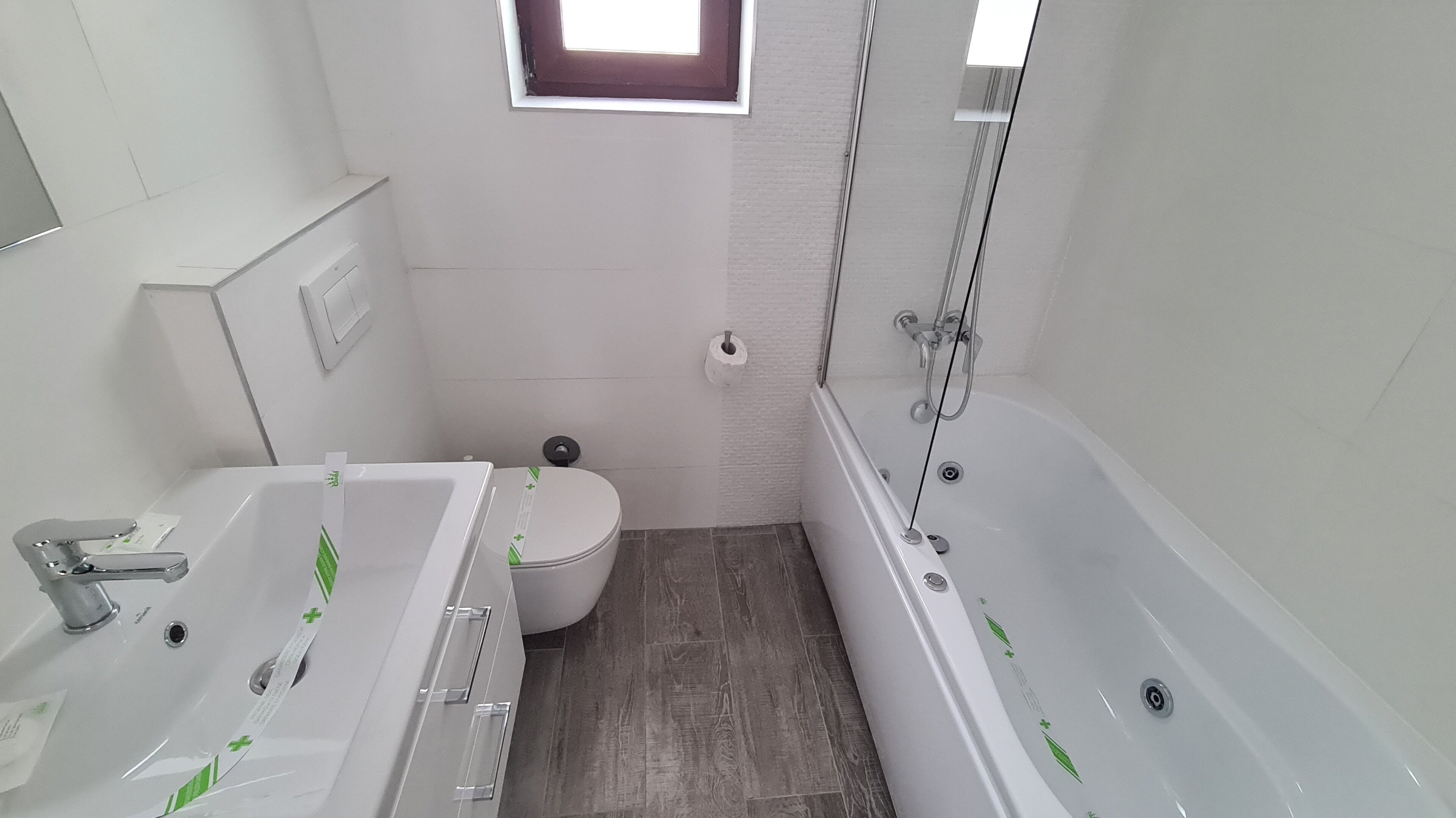 Een badkamer met een wit wastafelmeubel, toilet en bad, een raam boven het toilet en een douchehoek.