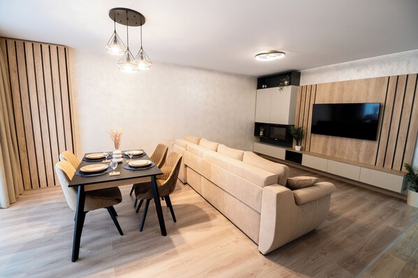 Ein modernes Wohnzimmer mit Essbereich, einem beige-farbenen Sofa, einem flachbildfernseher an der Wand und einer Wandpaneinung aus Holz.
