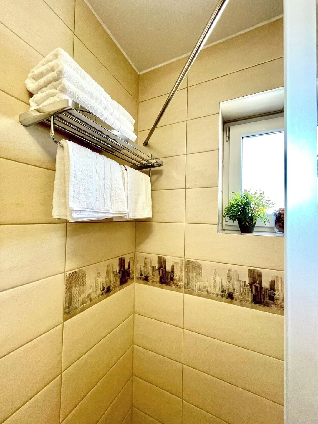 A corner bathroom with beige tiles, a towel rack with folded towels, a window with a plant, and a cityscape tile design.