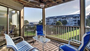 A covered outdoor area with a glass door leading to a balcony, furnished with chairs and a table, overlooking a residential area and a body of water.