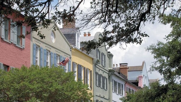 A row of colorful buildings with shutters, a flag, and trees in the foreground.