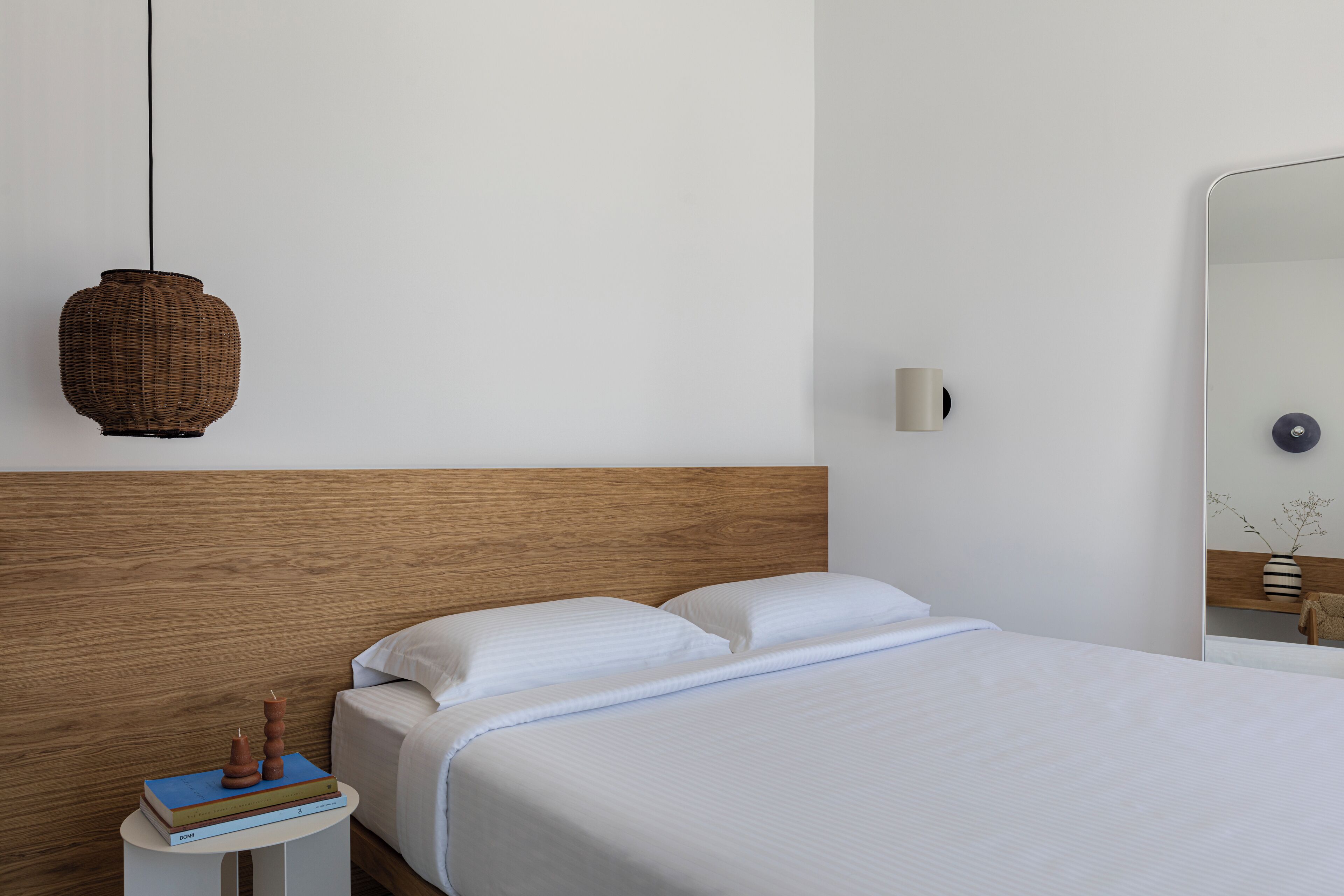 A neatly made bed with white linens, a wooden headboard, a small bedside table with books, a woven hanging light, and a wall-mounted mirror.