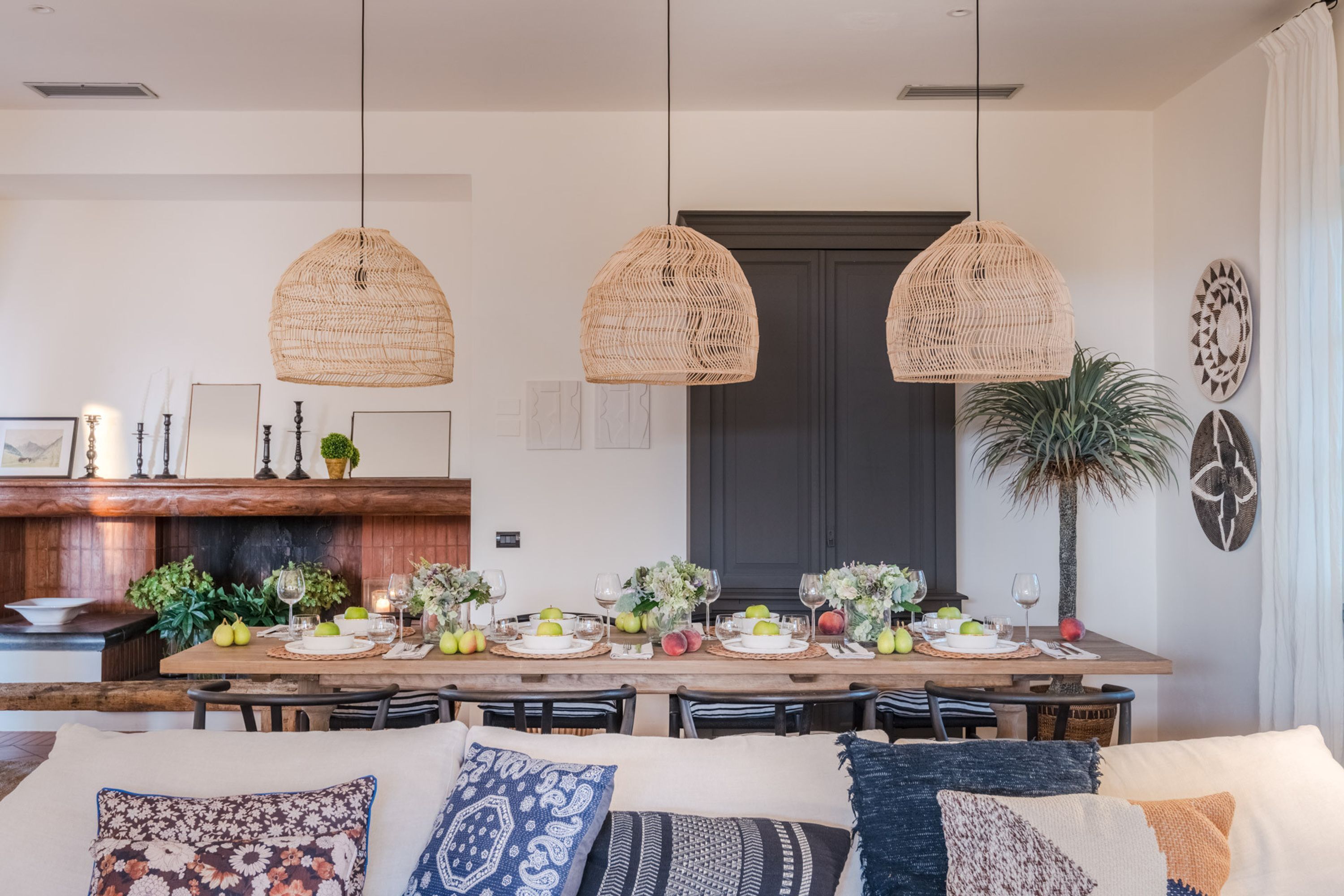 Uma sala de jantar com uma mesa comprida posta para seis pessoas, rodeada por cadeiras. Há luminárias trançadas penduradas sobre a mesa.