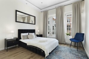 A modern bedroom with a black headboard, white bedding, a bedside table with a lamp, and a view of buildings through large windows.