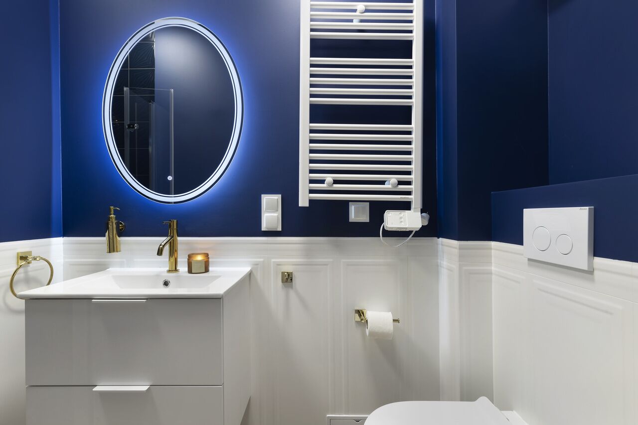 Un baño moderno con un lavamanos blanco, grifos dorados, un espejo redondo y un toallero blanco.