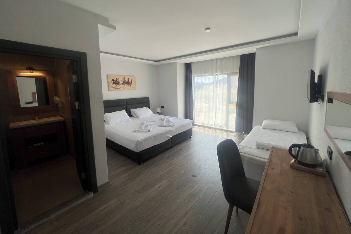 A hotel room with a large bed, a smaller bed, a desk with a kettle, a chair, a bathroom with a mirror, and a painting on the wall.