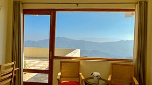 Ein Zimmer mit einem großen Fenster und Blick auf die Berge, zwei Korbstühle mit roten Kissen sowie ein kleiner Tisch mit einer Vase.