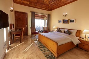 A bedroom with a wooden bed, a desk, and chairs, a balcony with curtains, and framed pictures on the wall. - Sumer hotel (Pachhad)