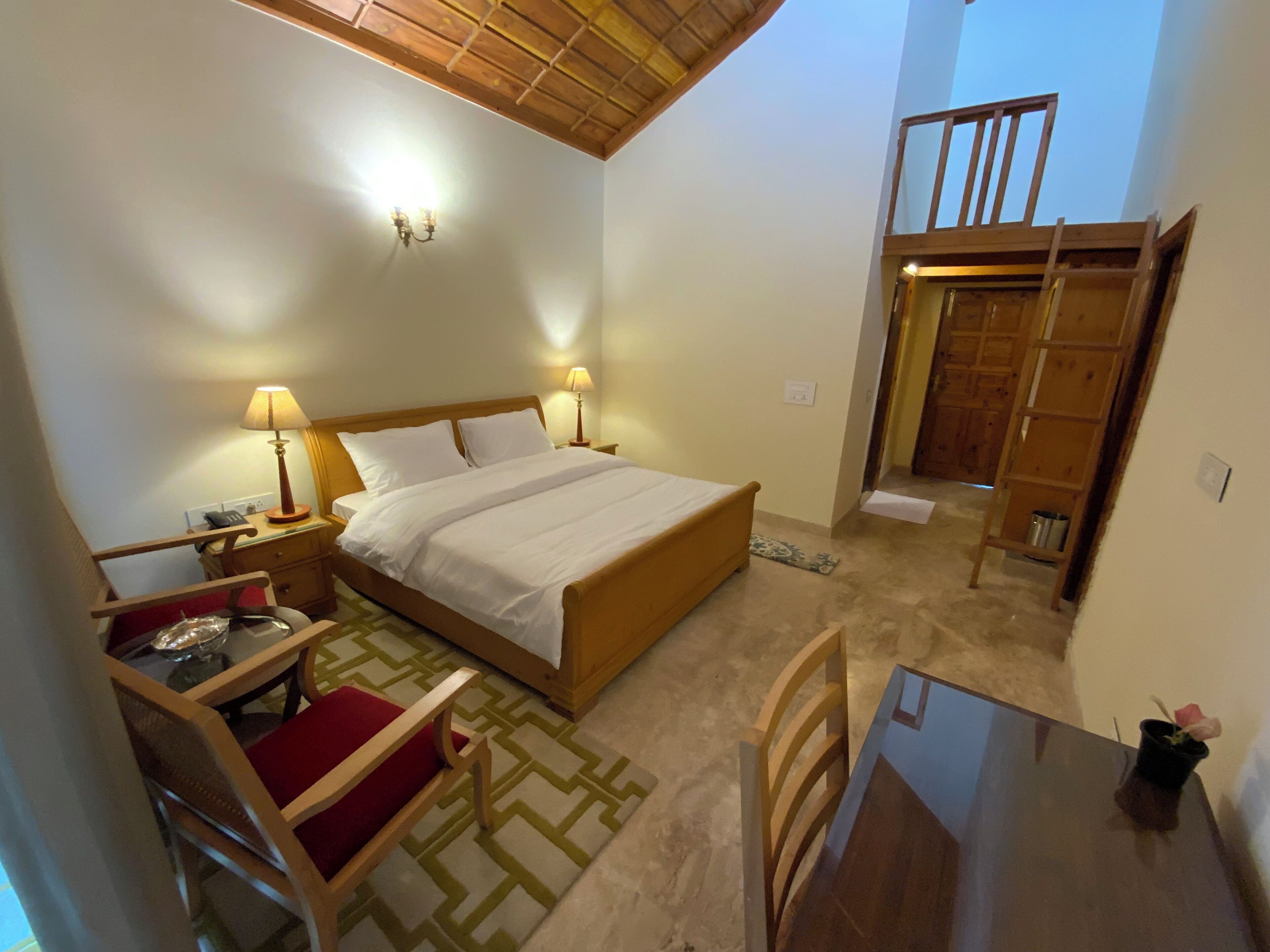 A bedroom with a wooden bed, bedside lamps, a small table, and a chair. There is a balcony with a wooden railing and a view of the sky.