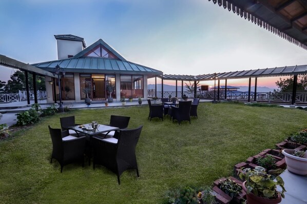 An outdoor patio area with a dining table and chairs, surrounded by a well-maintained lawn and potted plants.