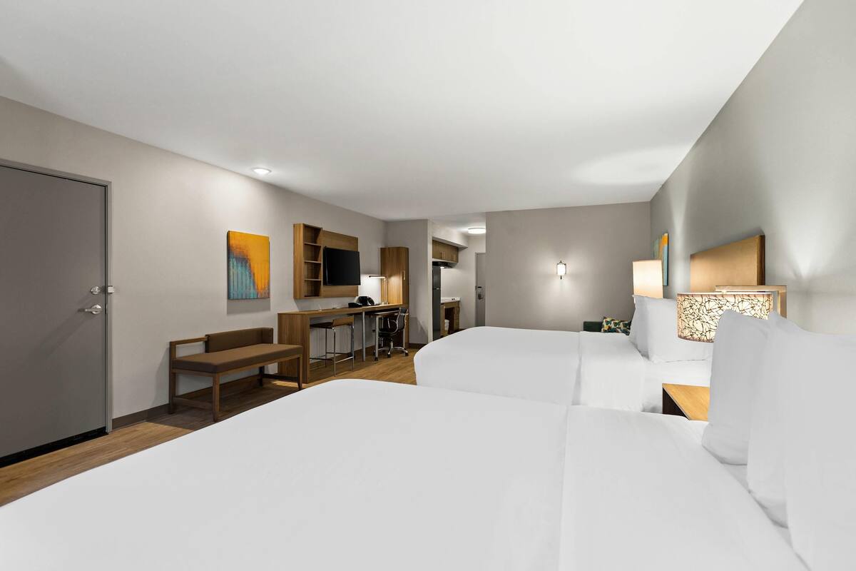 A hotel room with two beds, a desk, a chair, a bench, a TV, and a lamp.