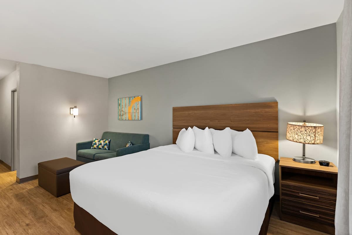 A hotel room with a large bed, a wooden headboard, a nightstand with a lamp, a blue armchair, and a small table with a lamp.