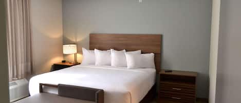 A hotel room with a large bed, a bedside table with a lamp, a chair, and a wooden dresser.
