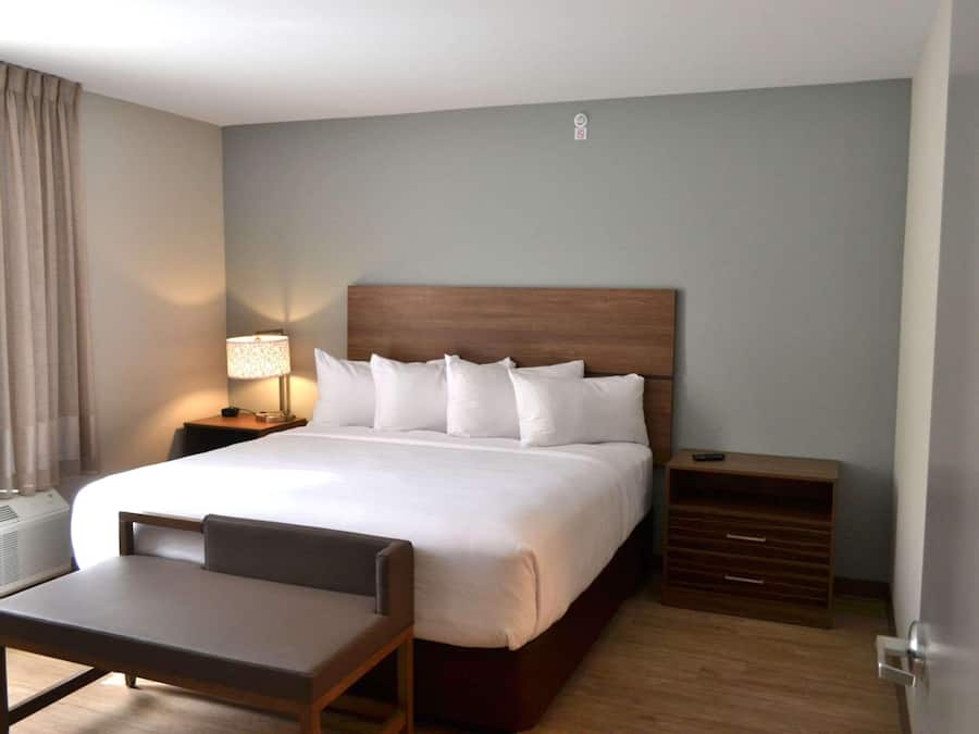 Quarto de hotel com uma cama grande, mesa de cabeceira com abajur, uma cadeira e cômoda de madeira.