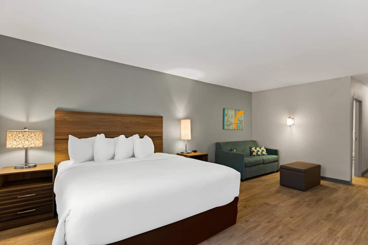 A hotel room with a large bed, a wooden headboard, two bedside tables with lamps, a green sofa, and a small ottoman.