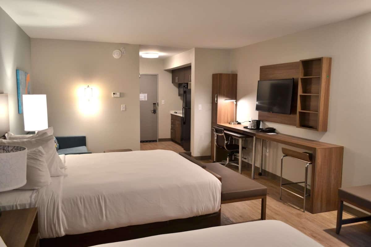 A hotel room with two beds, a desk with a chair, a television mounted on the wall, and a kitchenette area.
