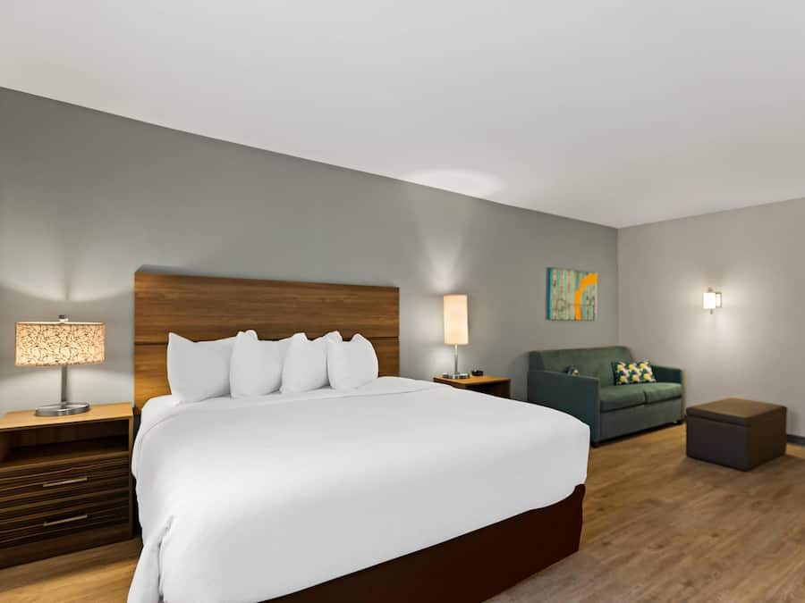Habitación de hotel con una cama grande, un cabecero de madera, dos mesitas de noche con lámparas, un sofá verde y un pequeño reposapiés.