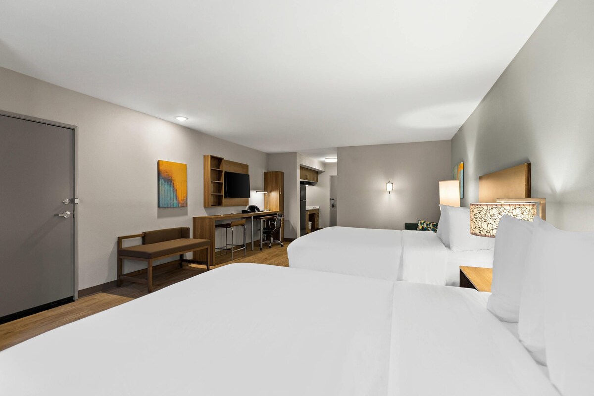 A hotel room with two beds, a desk, a chair, a bench, a TV, and a lamp.