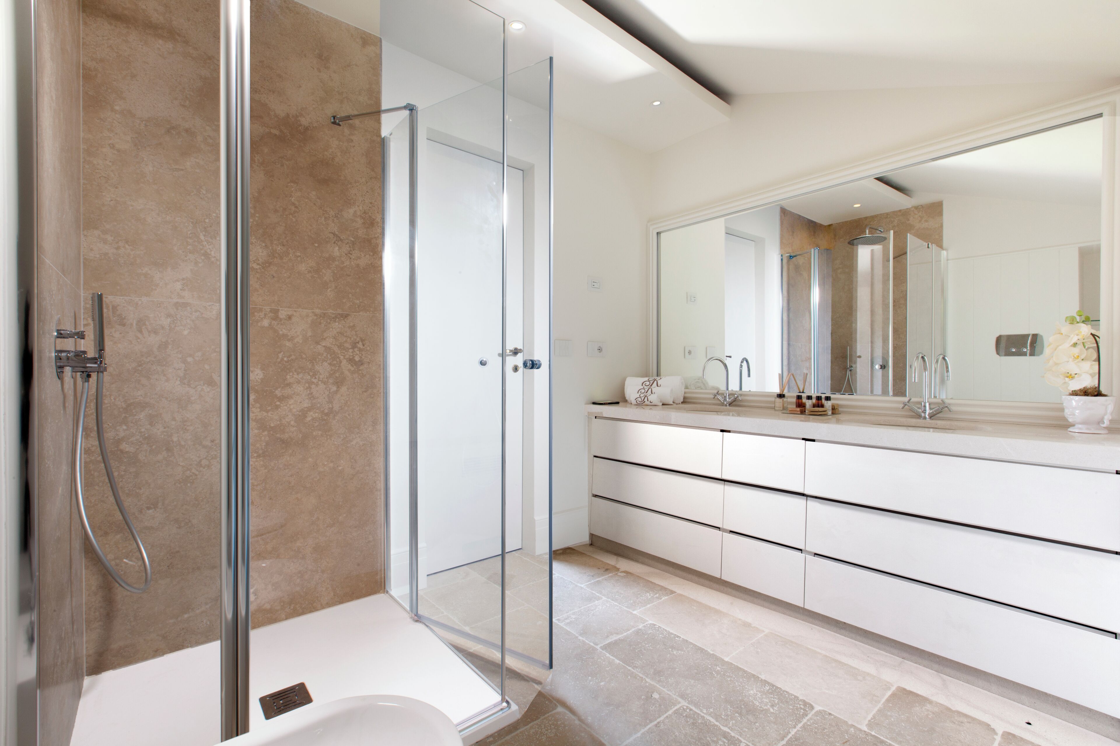 A modern bathroom with a glass-enclosed shower, a white vanity with a large mirror, and a beige tiled floor.