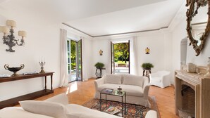 A spacious living room with a fireplace, white sofas, a coffee table, and a view of a garden through open doors.