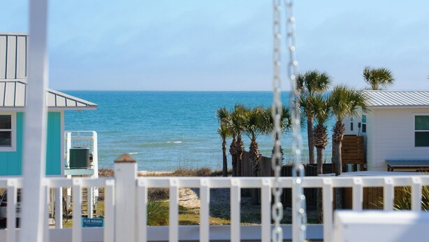 A beach view with a swing set, palm trees, and a building with a white roof. - Welcome to Sunset Sails & Wagging Tails! (St. George Island)