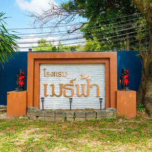 A sign with Thai script, flanked by two statues, set against a blue wall with a tree in the background. - Mayfa Hotel (Phuket)