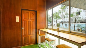 Une pièce avec une porte en bois, une fenêtre donnant sur des arbres et des immeubles, et une petite table en bois accompagnée de deux tabourets.