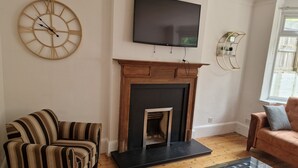Una sala de estar con chimenea, un reloj de pared grande, un televisor de pantalla plana y sillones a rayas.