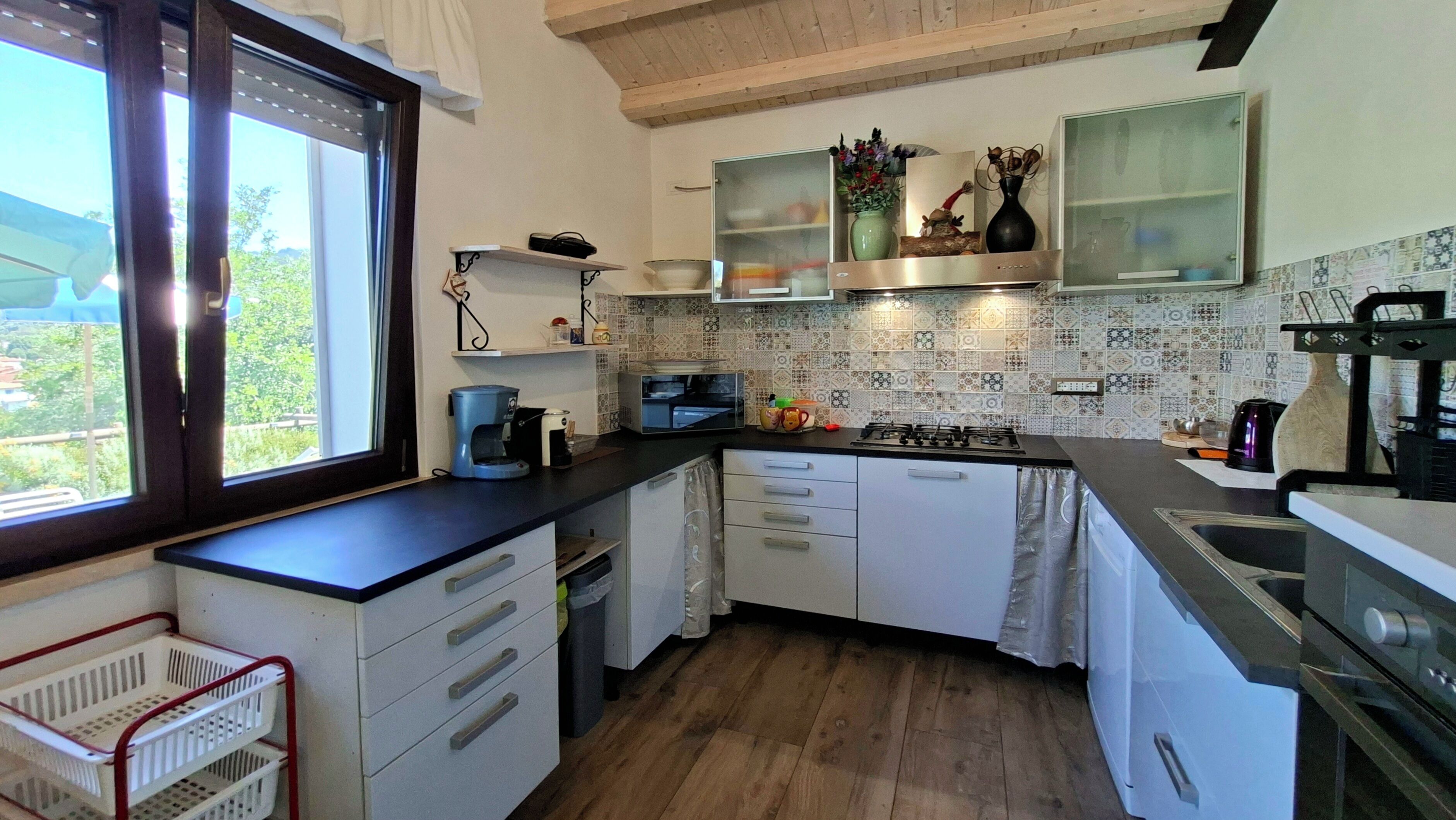 Une cuisine compacte avec des meubles blancs, une cuisinière à gaz, une machine à café et une fenêtre donnant sur un paysage verdoyant.