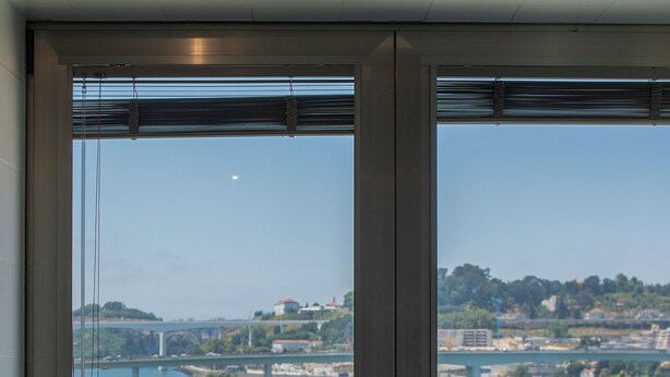A room with a glass table and a chair, a large window with a view of a river and a bridge, and a door with a handle. - Liiiving - Douro Riverside Apartment (Gondomar)