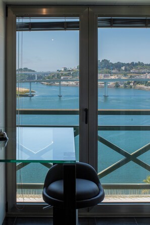 Een kamer met een glazen tafel en een stoel, een groot raam met uitzicht op een rivier en een brug, en een deur met een handvat.