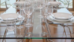 A dining table set with clear glassware and plates, positioned in a room with large windows overlooking a body of water. - Liiiving - Douro Riverside Apartment (Gondomar)
