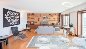 A modern living room with a sofa, dining table set, and a wall-mounted display of decorative items. - Liiiving - Douro Riverside Apartment (Gondomar)