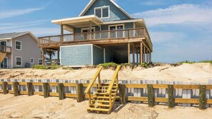 Una casa de playa de dos pisos con un balcón y una escalera de madera que lleva a la entrada.