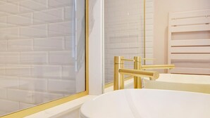 A bathroom with a white sink, gold fixtures, and a tiled wall. - A Stunning Studio-Loft in Place Vendôme - Louvre (Paris)