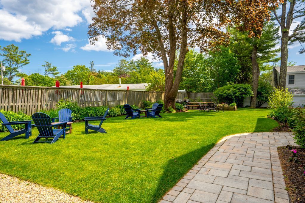 Un jardin avec un chemin pavé, une clôture en bois et un espace extérieur aménagé avec des chaises et une table.
