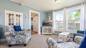 A living room with a blue and white floral sofa, a white side table, and a television on a white cabinet. There are large windows with curtains, a door leading to another room, and a ceiling light fixture. - Beautiful inn, Lighthouse Landing. #2 Sea Cliffs (Ogunquit)