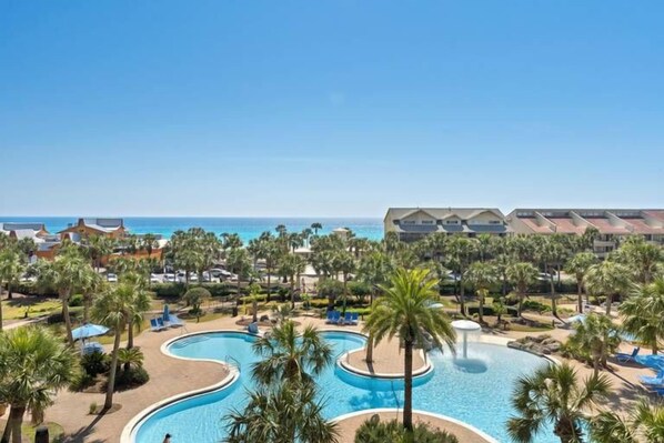 A resort with a large swimming pool surrounded by palm trees and lounge chairs, overlooking the ocean.