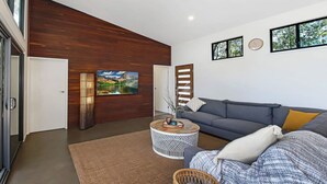 Uma sala de estar moderna com uma parede de destaque em madeira, um sofá cinzento, uma mesa de centro redonda e uma televisão de tela plana grande fixada na parede.