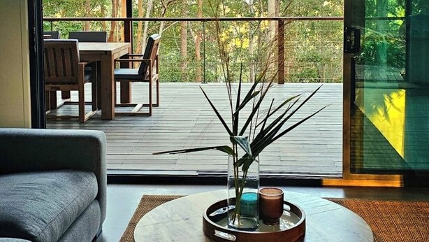 Uma sala de estar moderna com uma grande porta de vidro deslizante que dá para um deck externo, uma mesa de centro redonda com objetos decorativos e um sofá cinza.