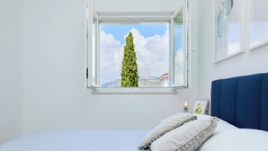 A bedroom with a bed, pillows, a window with a view of a tree and mountains, and a wall-mounted light fixture. - Hedera Estate, Hedera A35 (Dubrovnik)